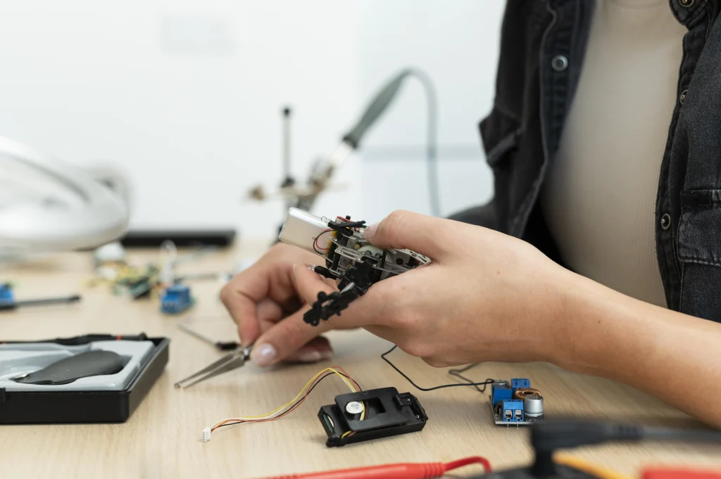 A person in a black shirt works on electronics, holding a small robot and using tweezers. Circuit boards and wires are scattered on a wooden table.
